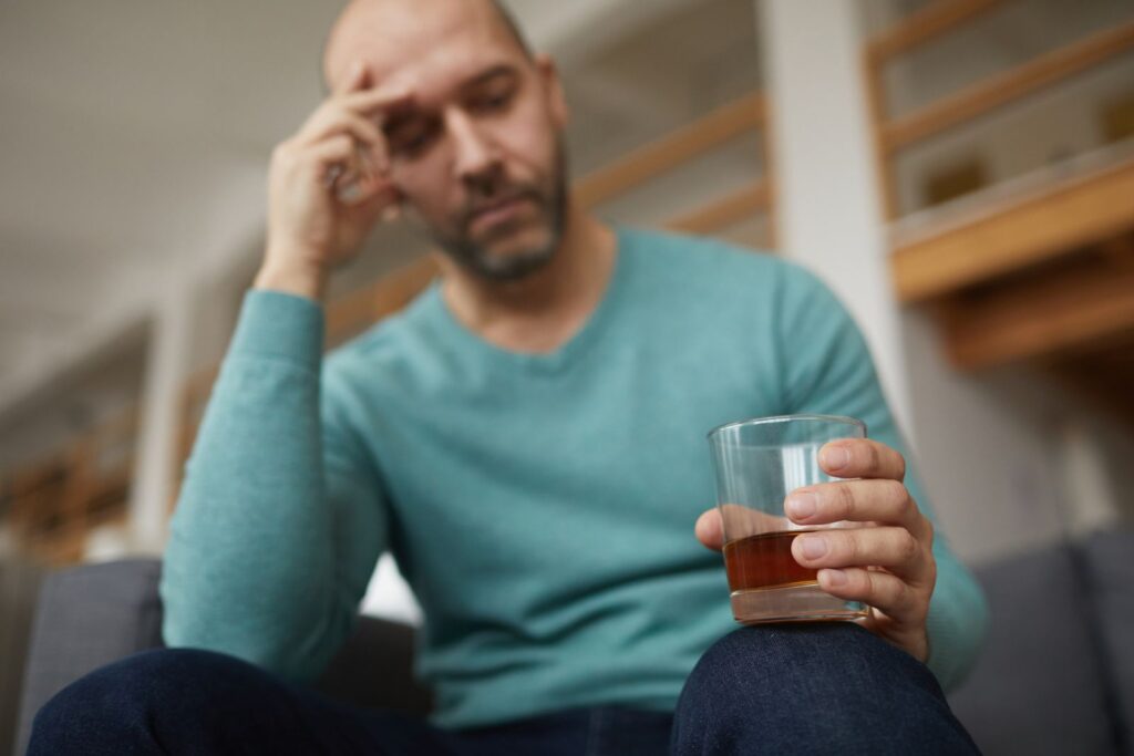 A man sits on a couch, holding a glass of alcohol, reflecting on the challenges of alcohol withdrawal.